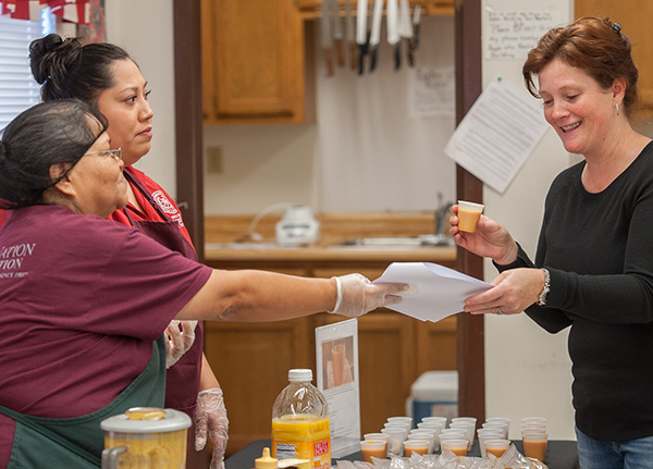 Two women wearing aprons and hairnets hand a customer a sample and a piece of paper